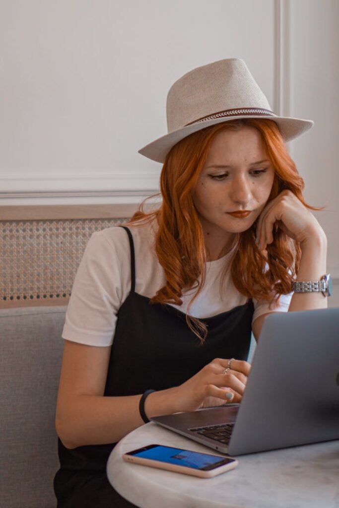 Redhead woman wearing a hat, focused on laptop work with a smartphone on the table.