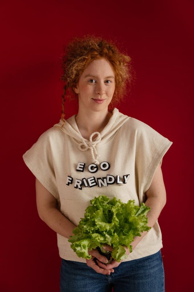 Red-headed woman holding lettuce against a red background wearing an 'eco-friendly' shirt.